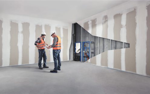 Two installers standing in an empty room and looking at a technical drawing one of them is holding. A cut-out in the adjacent wall reveals a complex plumbing system running through it. | Pipelife