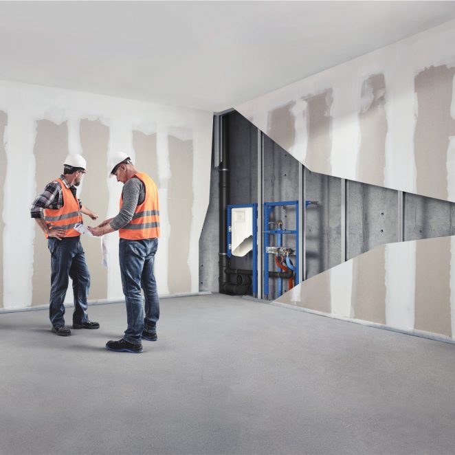 Two installers standing in an empty room and looking at a technical drawing one of them is holding. A cut-out in the adjacent wall reveals a complex plumbing system running through it. | Pipelife